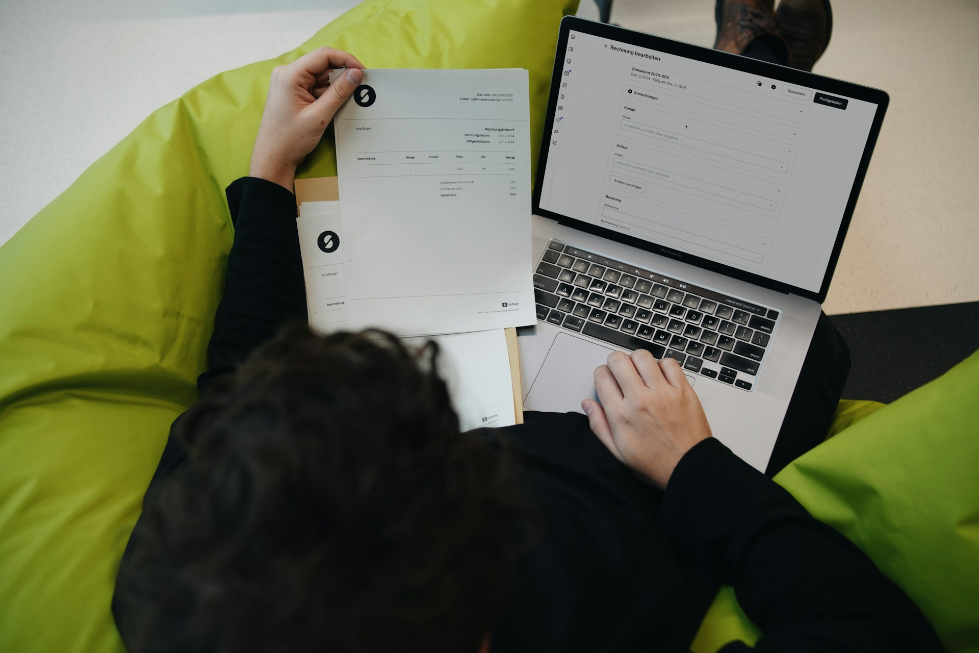 A person sits on a green beanbag holding a document in one hand and using a laptop with an online form open on the screen, appearing to review or enter information.